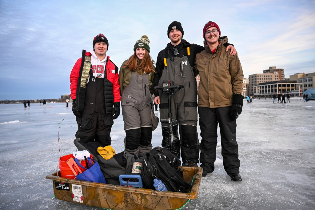 Four people wearing winter clothing and holding ice fishing gear smile for the camera as they stand on a frozen lake.