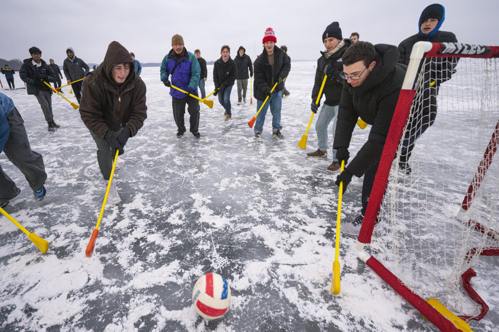 Several students holding brooms chase after a ball on an icy lake.