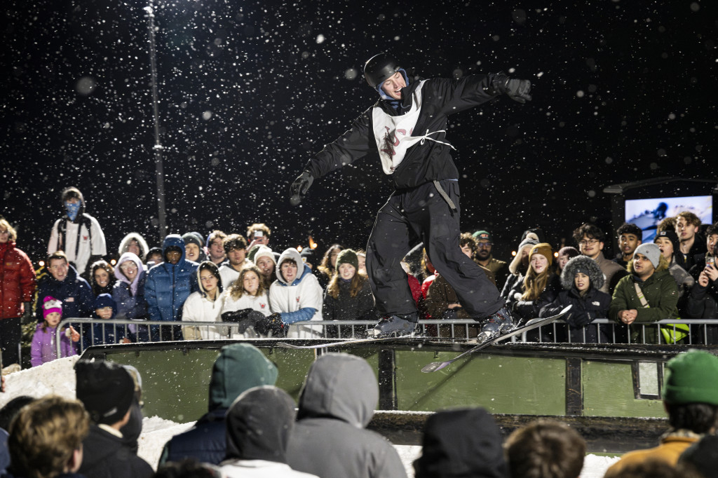 A skier flies through the air as his skies slide on a rail, with the night sky behind him and snowflakes around him.